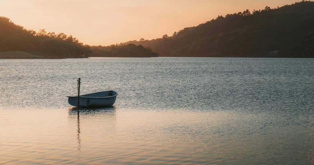 A small pale rowing boat moored to a post on still lake water at sunset, with wooded hills on the far shore